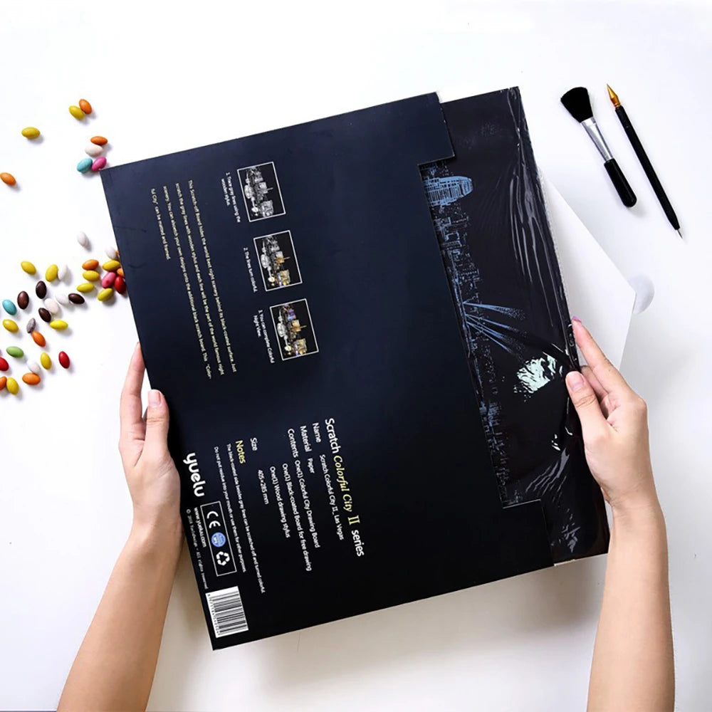 A black scratch art kit featuring a city night scene, held by a person against a white background.