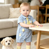 A baby boy wearing a blue short-sleeve romper with bear and mushroom appliqués sitting on the floor.