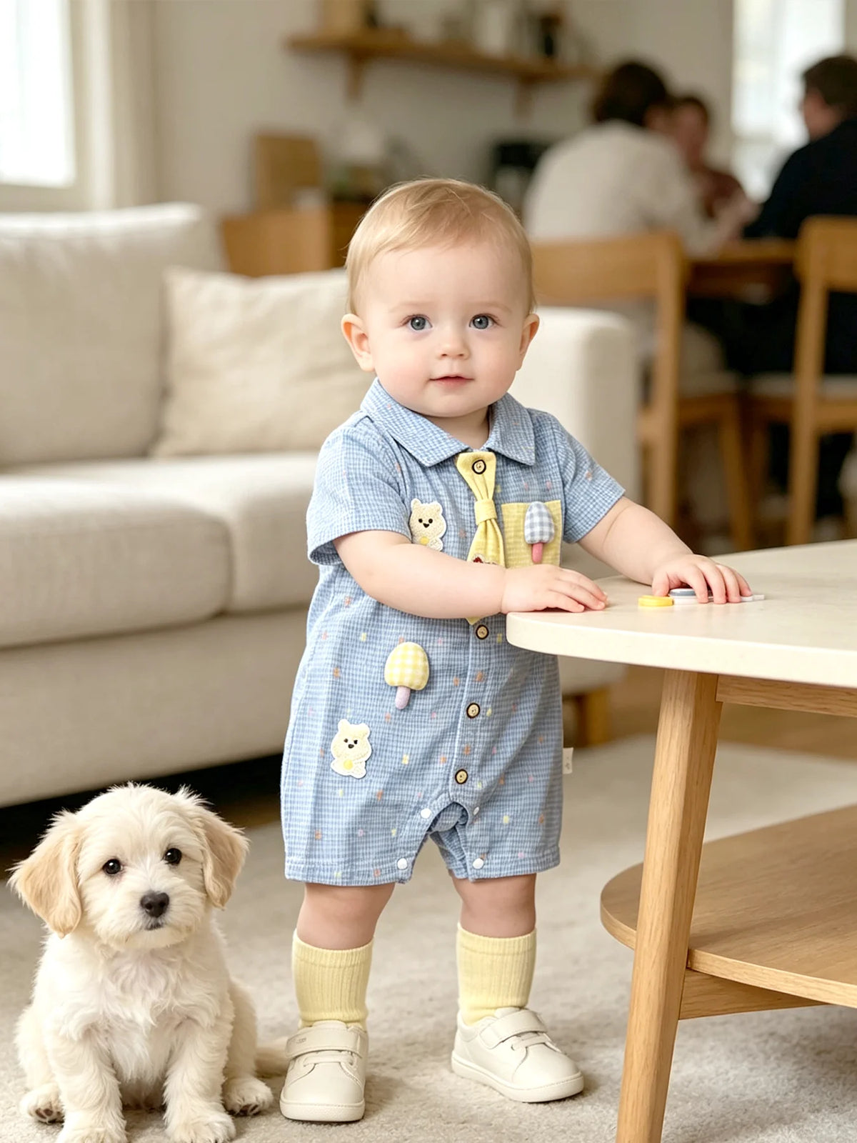 A baby boy wearing a blue short-sleeve romper with bear and mushroom appliqués sitting on the floor.