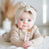 Un bébé porte un pull tricoté beige et tient un hochet éducatif Arc-en-ciel Montessori.