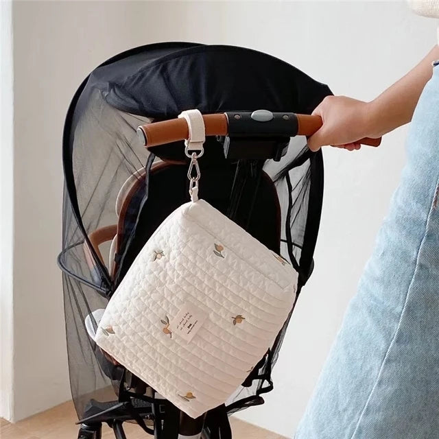 A hand is placing a quilted white diaper bag with embroidery onto a black baby carrier or stroller.