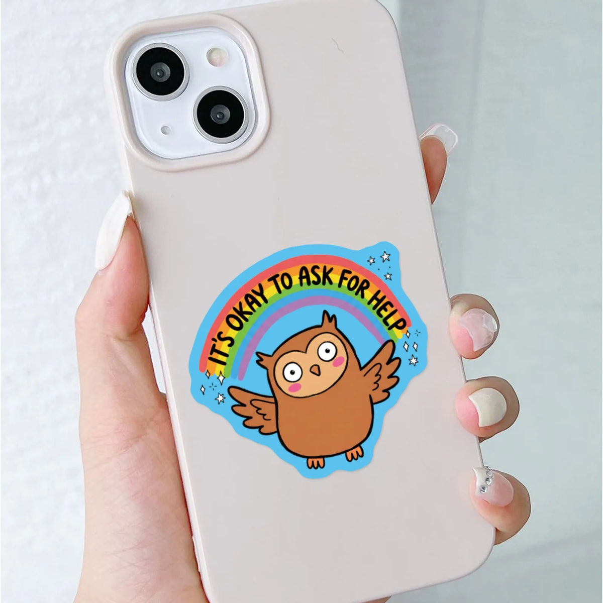A hand holds a white phone case featuring an owl sticker with the text It's okay to ask for help surrounded by a rainbow.