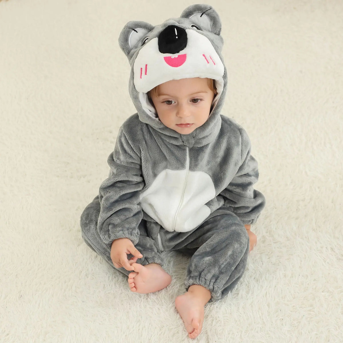 A baby wearing a gray animal hooded romper with black facial features sitting on a white fluffy surface.