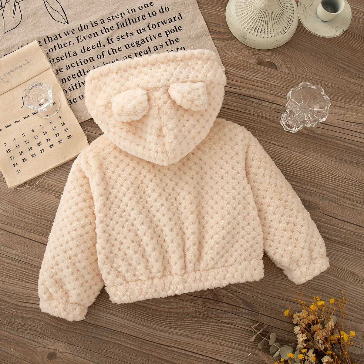 A cream-colored, knitted plush hooded winter jacket for a baby girl is displayed on a wooden surface.