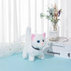 A white plush cat toy with pink ears and blue eyes sitting on a light blue surface next to a glass vase of dried flowers.