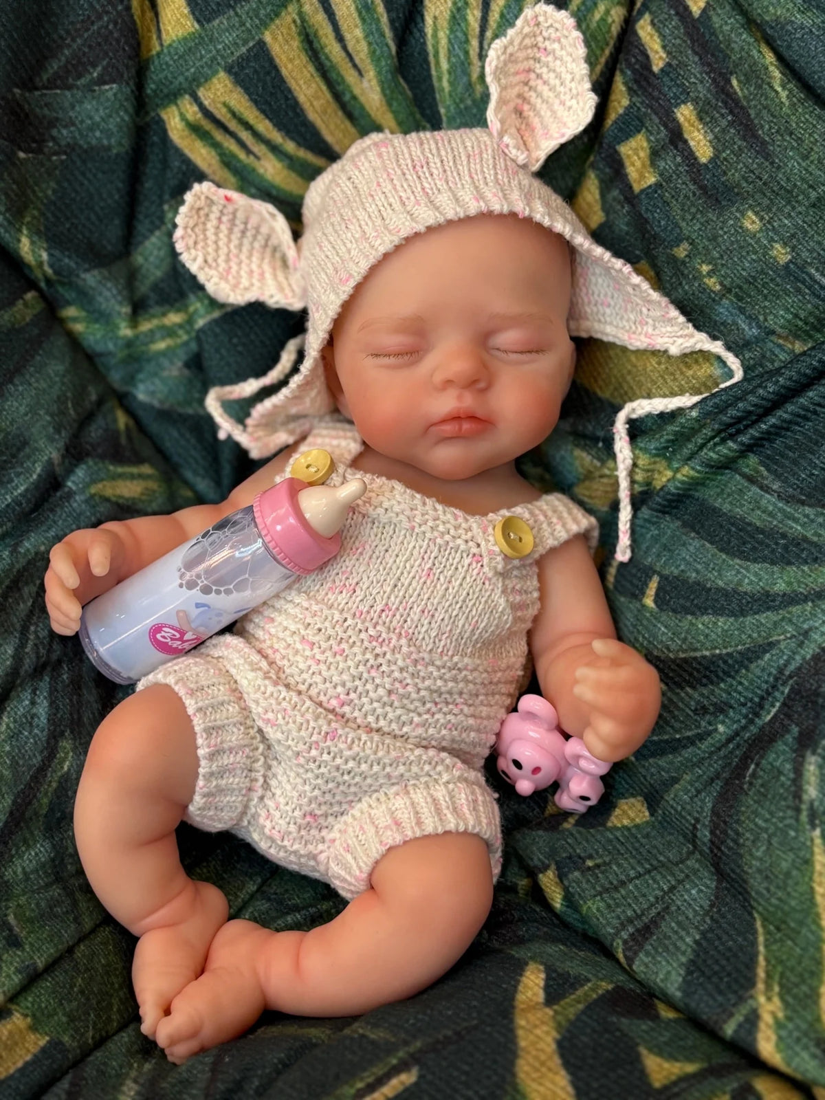 A hand-painted silicone reborn doll with pink ears and a light pink knitted outfit lying on green fabric.