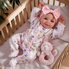 A baby doll wearing a pink bow and a floral outfit is lying on a light pink surface.