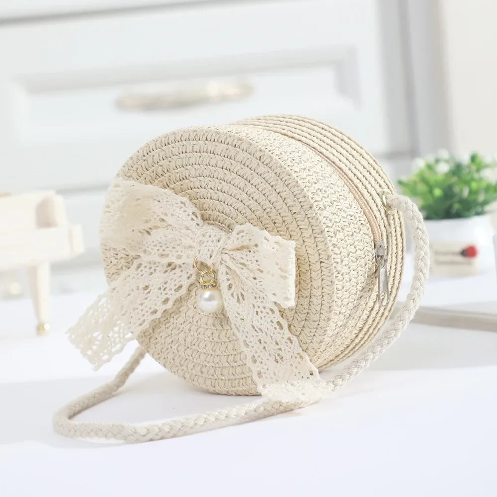 A beige handwoven straw crossbody bag featuring a delicate white lace bow and pearl detail.