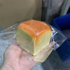 Une main tient un pain décoratif carré, doux, avec des couleurs jaune et orange dans un emballage en plastique transparent.