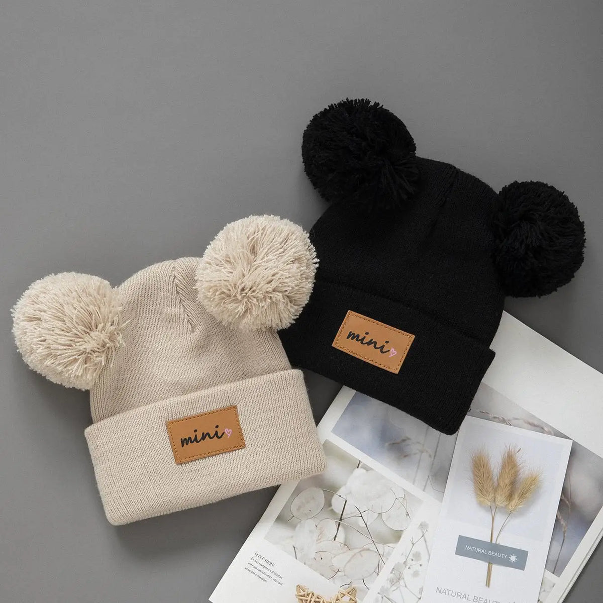 A black knit baby beanie hat with black and cream faux fur pom-poms and a mini label.