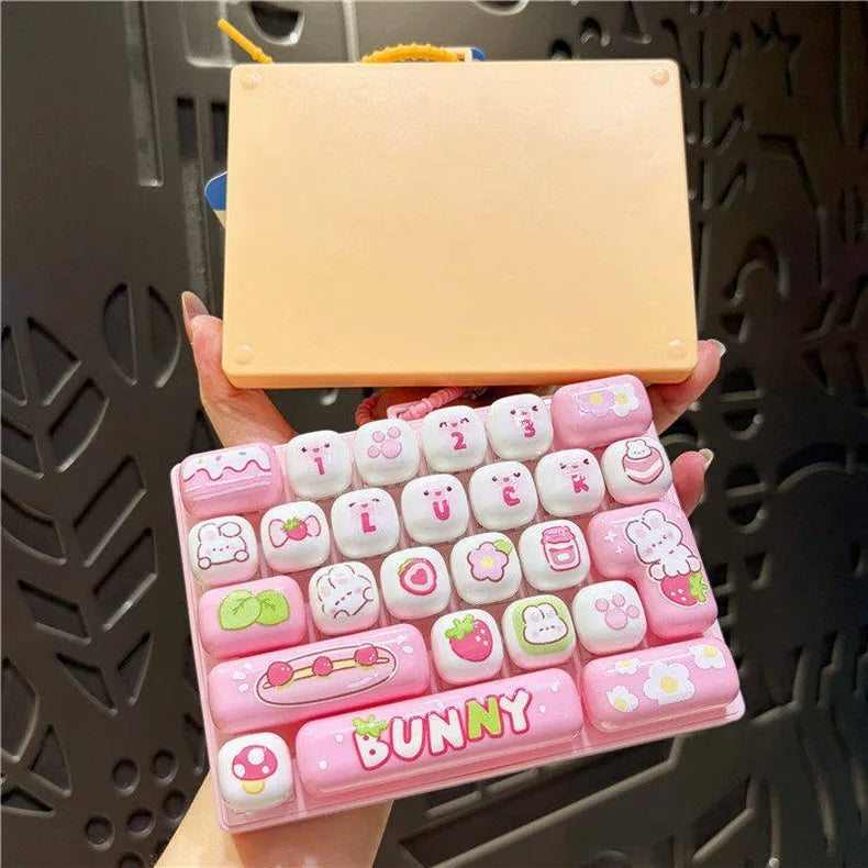 A pink and white set of 26 keycap charms shaped like various cartoon animals is held above a square beige item.