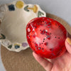 A hand holds a glossy red stress ball decorated with small stars and hearts, resting on a beige ceramic dish.