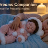A sleeping baby doll wearing a pink and white fluffy winter romper is resting on a blue blanket in a soft, dimly lit bedroom.