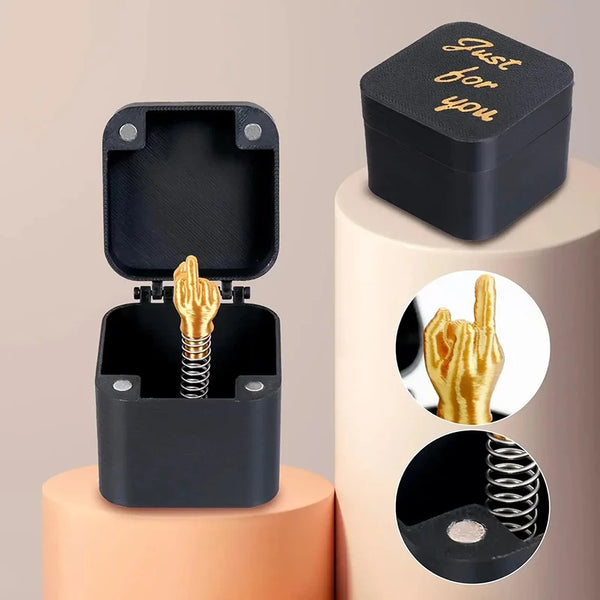 A black pop-up gift box with a gold middle finger hand visible inside, displayed on a light pink stand.