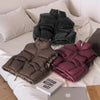 A stack of three padded down vests in shades of gray, brown, and magenta resting on a white bed with pillows.