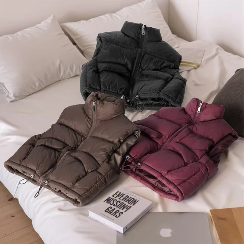 A stack of three padded down vests in shades of gray, brown, and magenta resting on a white bed with pillows.