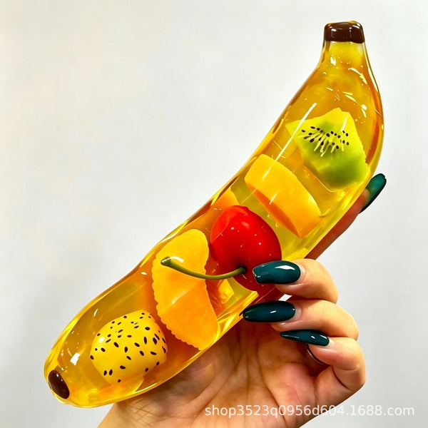 Une main tient un jouet squishy jaune en forme de banane, rempli de morceaux de fruits colorés.