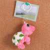 A pink and green turtle plush toy attached to a silver keyring charm on a brown cork board.