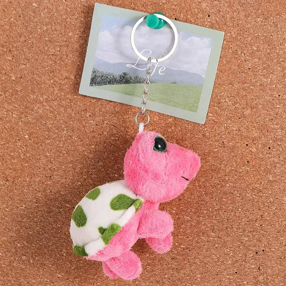 A pink and green turtle plush toy attached to a silver keyring charm on a brown cork board.