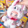 A white fluffy collectible doll with pink accents and a small white figure inside its packaging box.