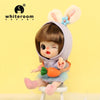 A small, light brown doll with pink hair and large ears is shown wearing a purple hood and an orange accessory.