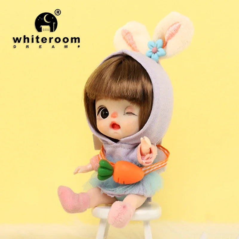 A small, light brown doll with pink hair and large ears is shown wearing a purple hood and an orange accessory.
