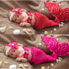 A newborn baby lying on brown fabric wearing a red knit mermaid tail skirt and a matching pearl headband.