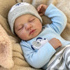 A sleeping newborn vinyl doll wearing a light blue long-sleeved shirt and a grey knitted hat, resting on a soft beige blanket