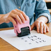 A person uses a pink roller stamp set to make math equations on a piece of paper at a wooden table.