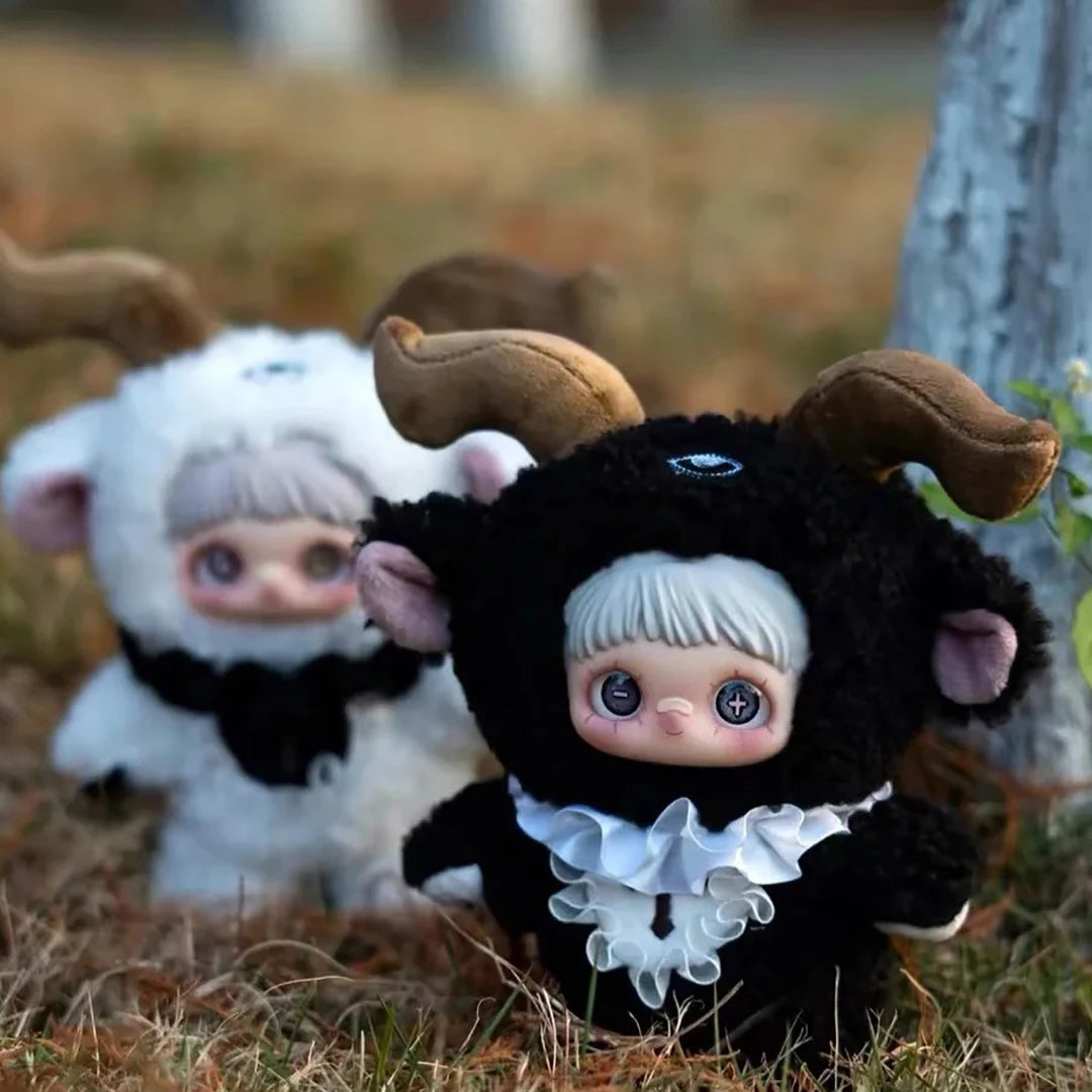 A black and white plush doll resembling a ram or goat with large ears and a ruffled collar sitting on dry grass.