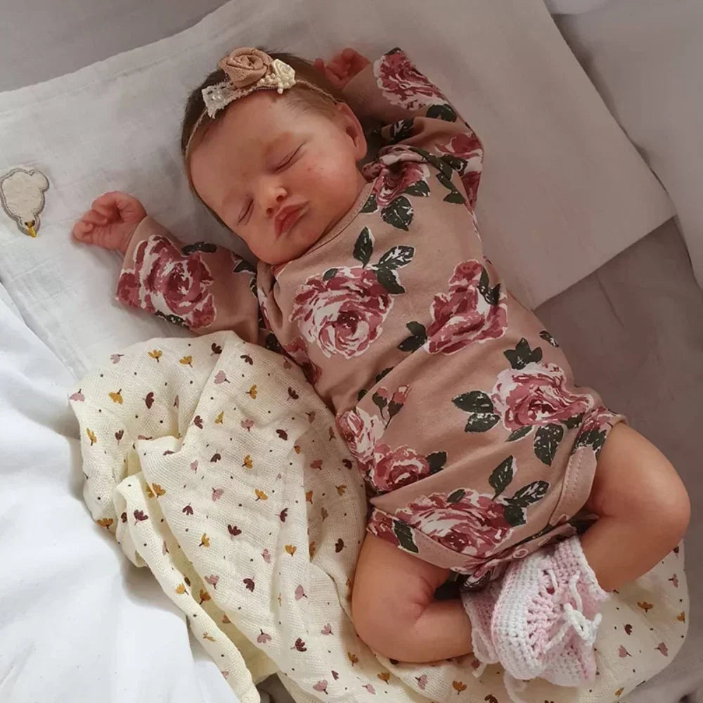 A newborn doll with soft silicone body is sleeping on white fabric, adorned with floral patterned clothing and a headband.