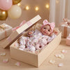 A newborn baby doll in pink clothing is displayed inside a wooden box with soft petals and ribbon.