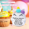 Weiße Häkel-Einhornpuppe mit farbenfrohen Ohren neben einem Cupcake und Text, der positive Energie wählt.