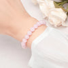 A close-up of a natural pink quartz stretch bracelet worn on a wrist, next to a white sleeve and a white rose.