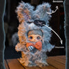 A fluffy gray plush doll with a face, orange bow, and detachable features, shown against a dark background.