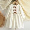 A cream-colored girls' long sleeve bow detail A-line dress hanging on a wooden hanger.