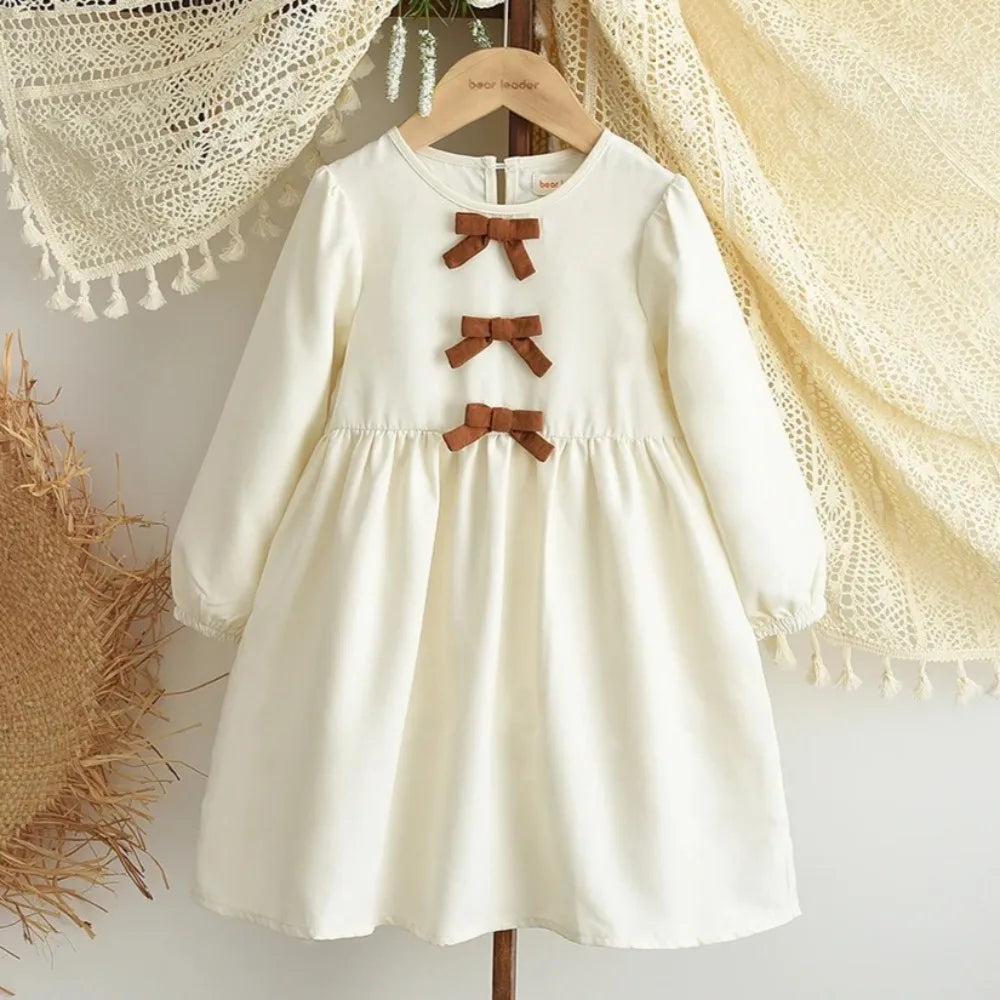 A cream-colored girls' long sleeve bow detail A-line dress hanging on a wooden hanger.