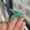A hand holds a turquoise and gold rhinestone turtle brooch pin against a blurred window background.