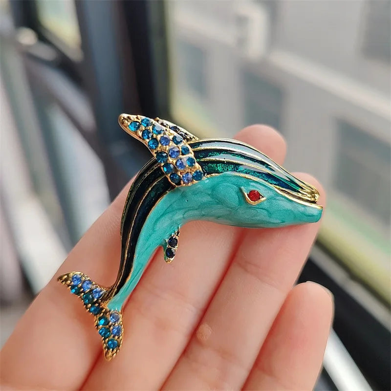 A hand holds a turquoise and gold rhinestone turtle brooch pin against a blurred window background.
