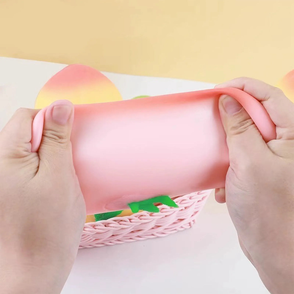 Hands squeezing a pink peach-shaped squishy stress relief toy inside a pink woven basket.