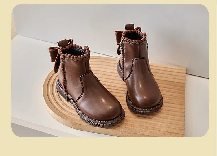 A pair of brown leather ankle boots with bow-knot detailing and a side zipper resting on a wooden surface.