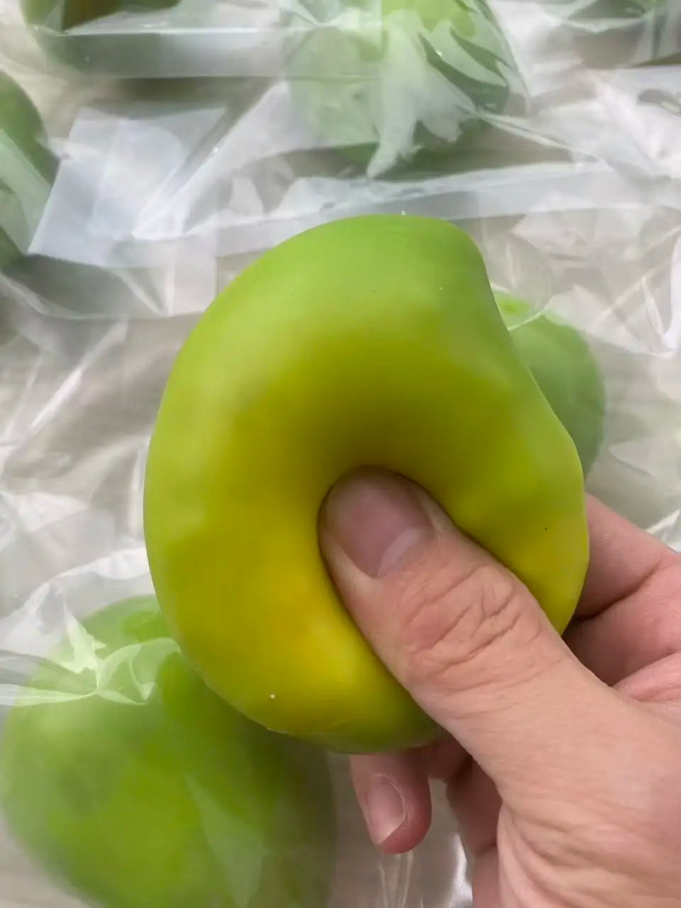 A hand holds a bright lime green, oval-shaped stress ball with a squishy texture inside a clear plastic bag.