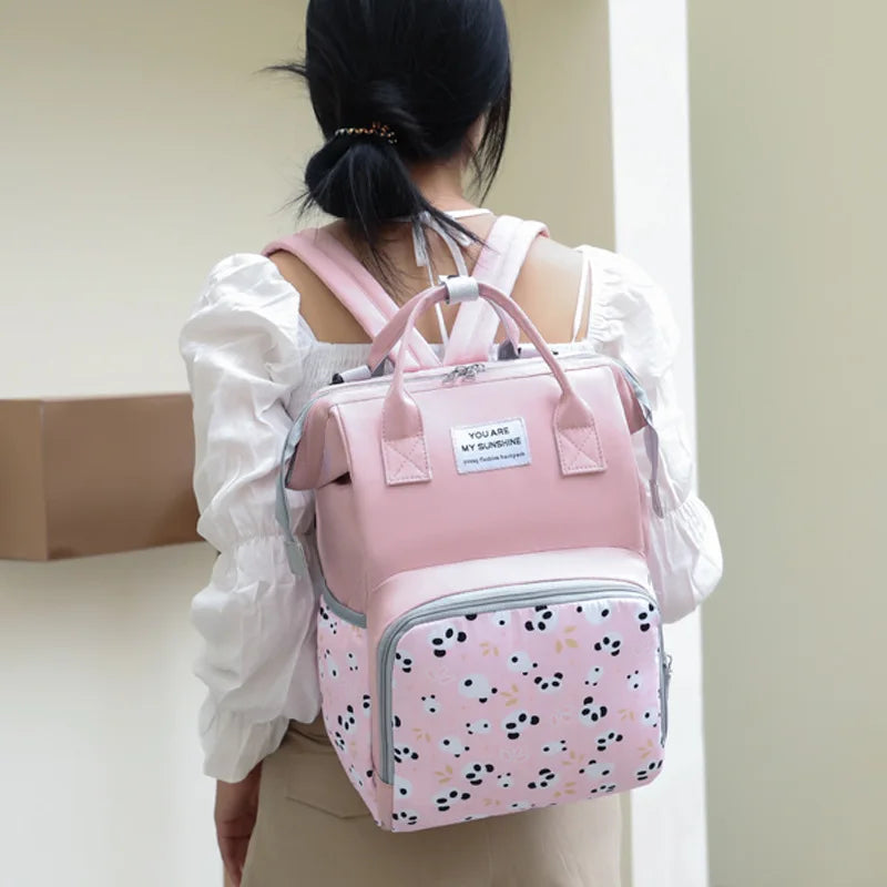 A woman is shown from the back carrying a pink Mommy backpack with a floral pattern.