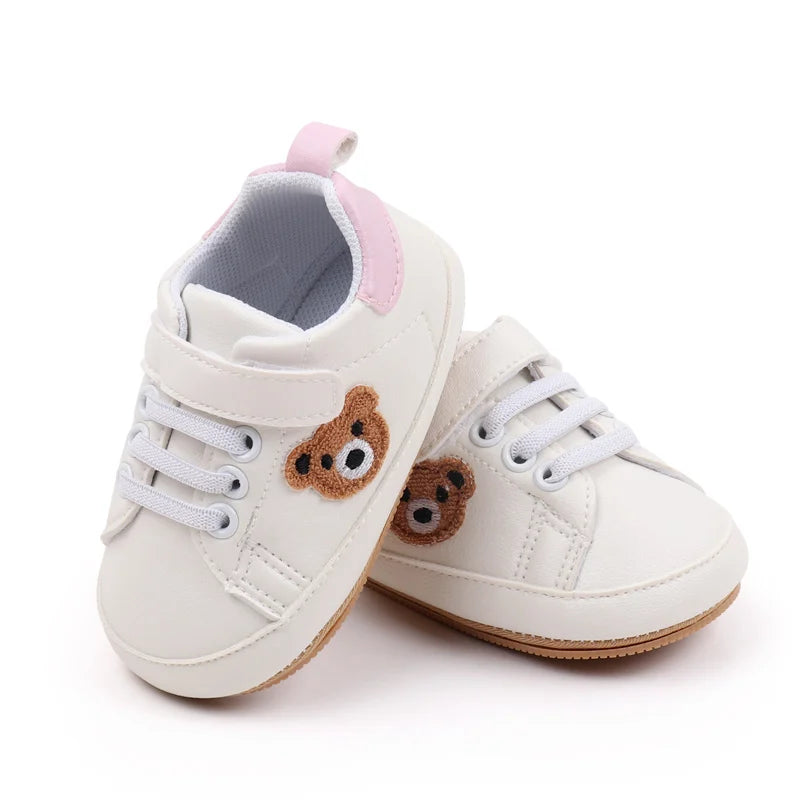 A pair of light beige baby sneakers with pink accents and a cute brown bear design.