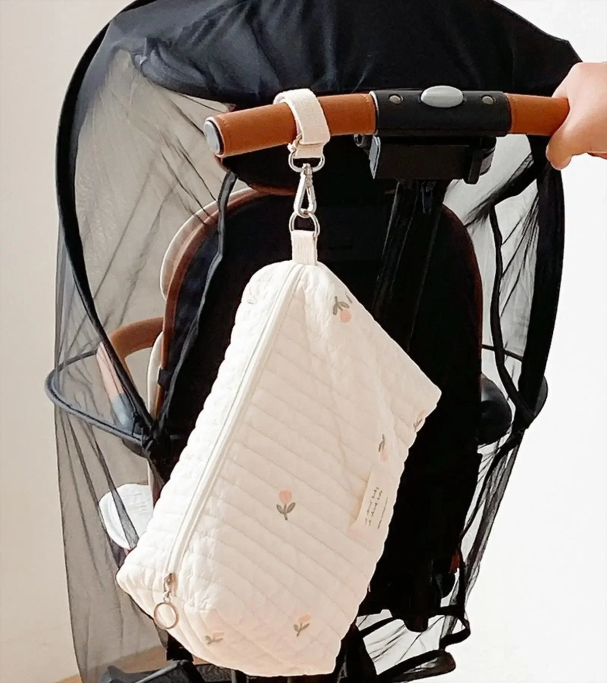 A quilted white baby diaper caddy organizer hanging from a brown leather strap on a black stroller.