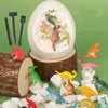 A dinosaur fossil dig kit showing a white eggshell containing small dinosaur fossils and various excavation tools on a green 