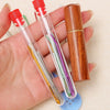 A hand holds a clear glass tube containing a set of stainless steel sewing needles with colored tips and a wooden storage tub