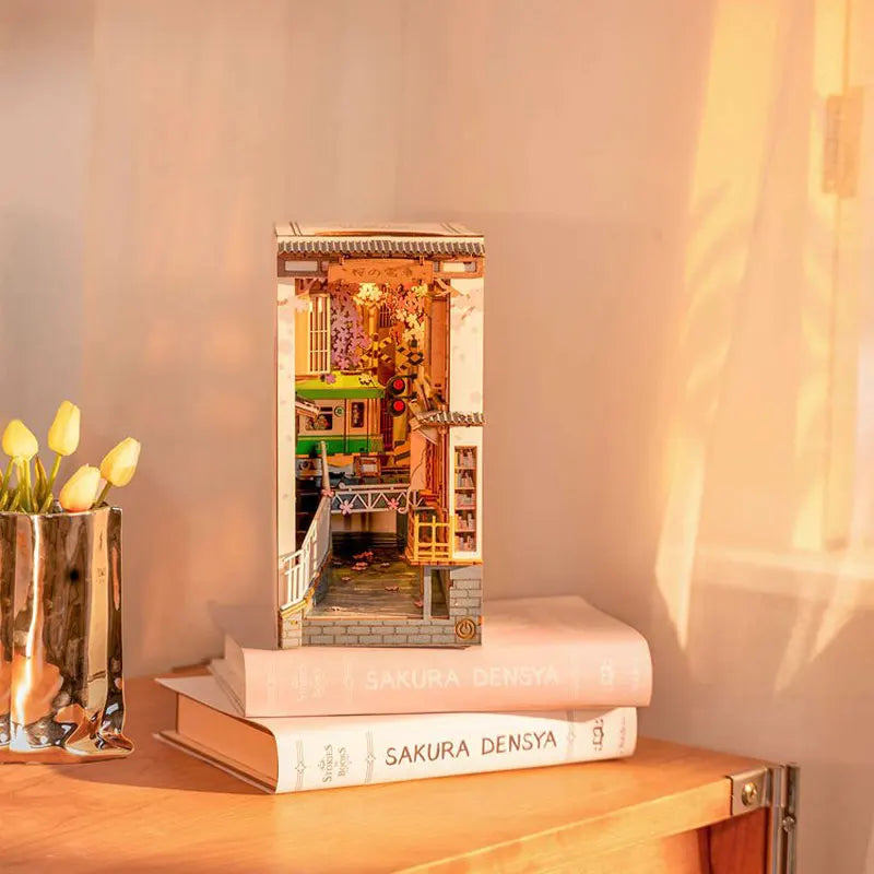 A miniature dollhouse kit displayed in a wooden frame with interior lighting on a light wood surface.