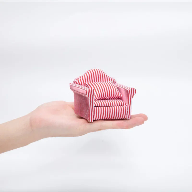 A hand holds a miniature red and white striped sofa and pillow set for a dollhouse.