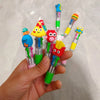 A hand holds several cute cartoon four-color ballpoint pens with colorful character toppers.